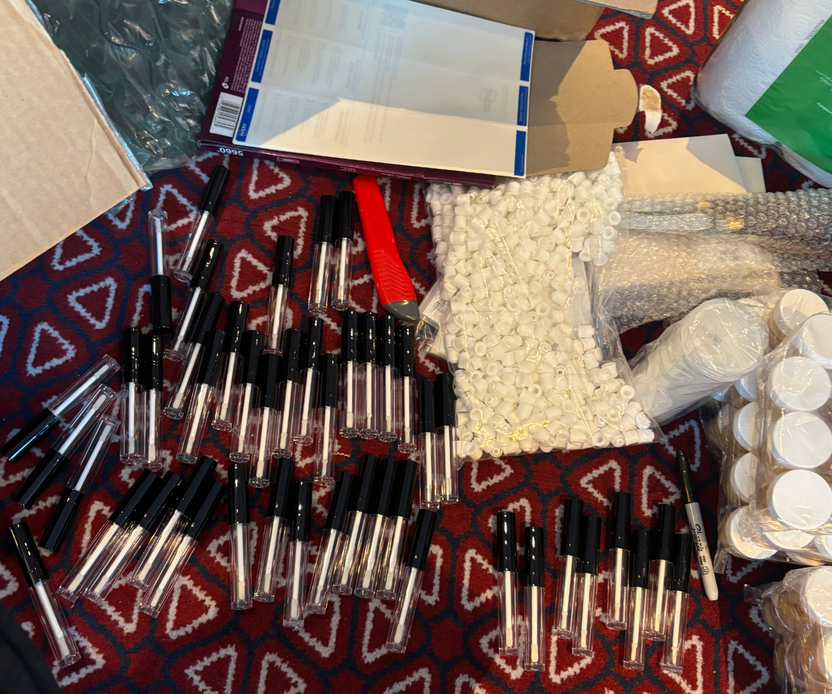 Overhead view of perfume-making supplies arranged on a patterned red textile, including small glass vials, pipettes, white containers, and cardboard boxes